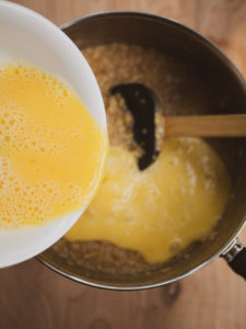 Anna's hand pouring mixed raw eggs into a sauce pot with cooked oatmeal for the savory oatmeal recipe