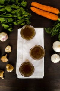 Beef Broth in 3 little mason jars on a white napkin with mushrooms on the left and garlic on the right.