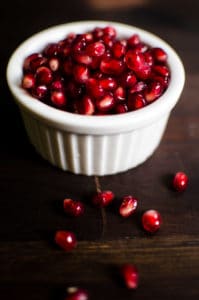 Close up look of pomegranate seeds in a white bowl on a dark wood background. Stories from garlicdelight.com.